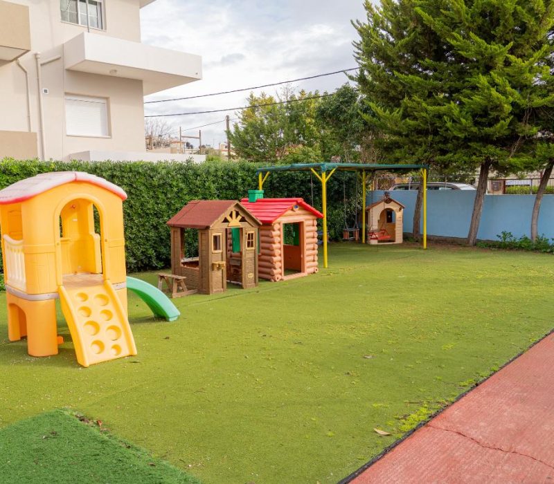 Εξωτερική αυλή παιδικού σταθμού στη Χίο με τάπητα ασφαλείας, τσουλήθρες και σπιτάκια παιχνιδιού.